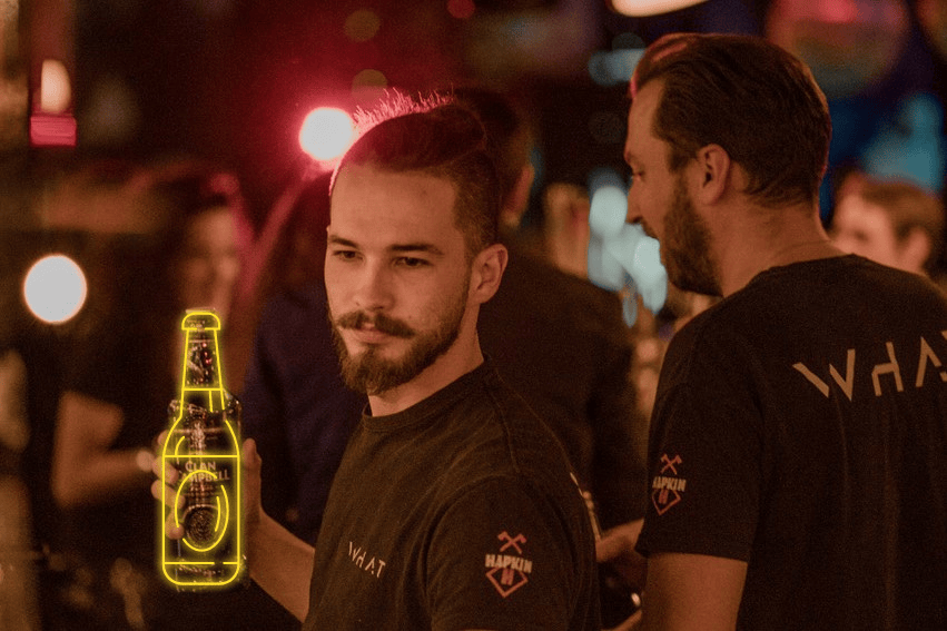 Laurent le barman avec sa moustache et ses cheuveux long entrain de travailler avec une bouteille d'alcool, avec son collègue dans une ambiance sombre